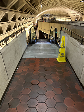 A picture of an escalator. There is a yellow wet floor cone slightly to the right of center of the image, to the right side of the escalator entrance. On both sides of the escalator there are half walls with railings on top. The background looks upon the escalator coming up from this platform.